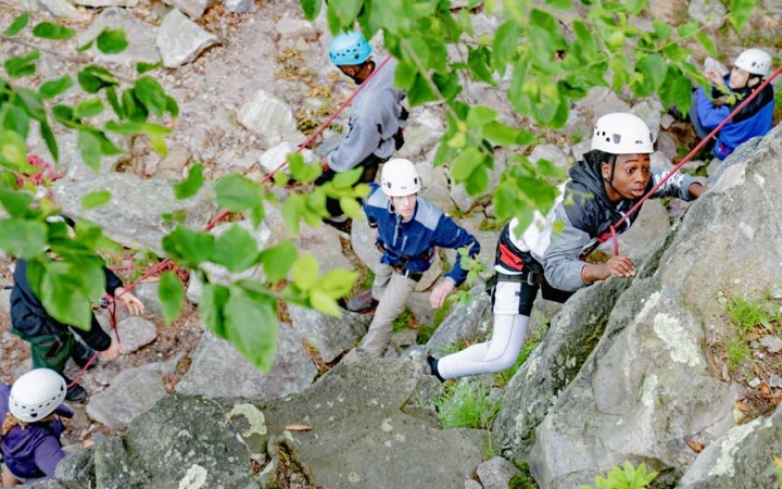 Looking down, you can see several people wearing helmets standing at the base of a rock wall. One person is climbing. 
